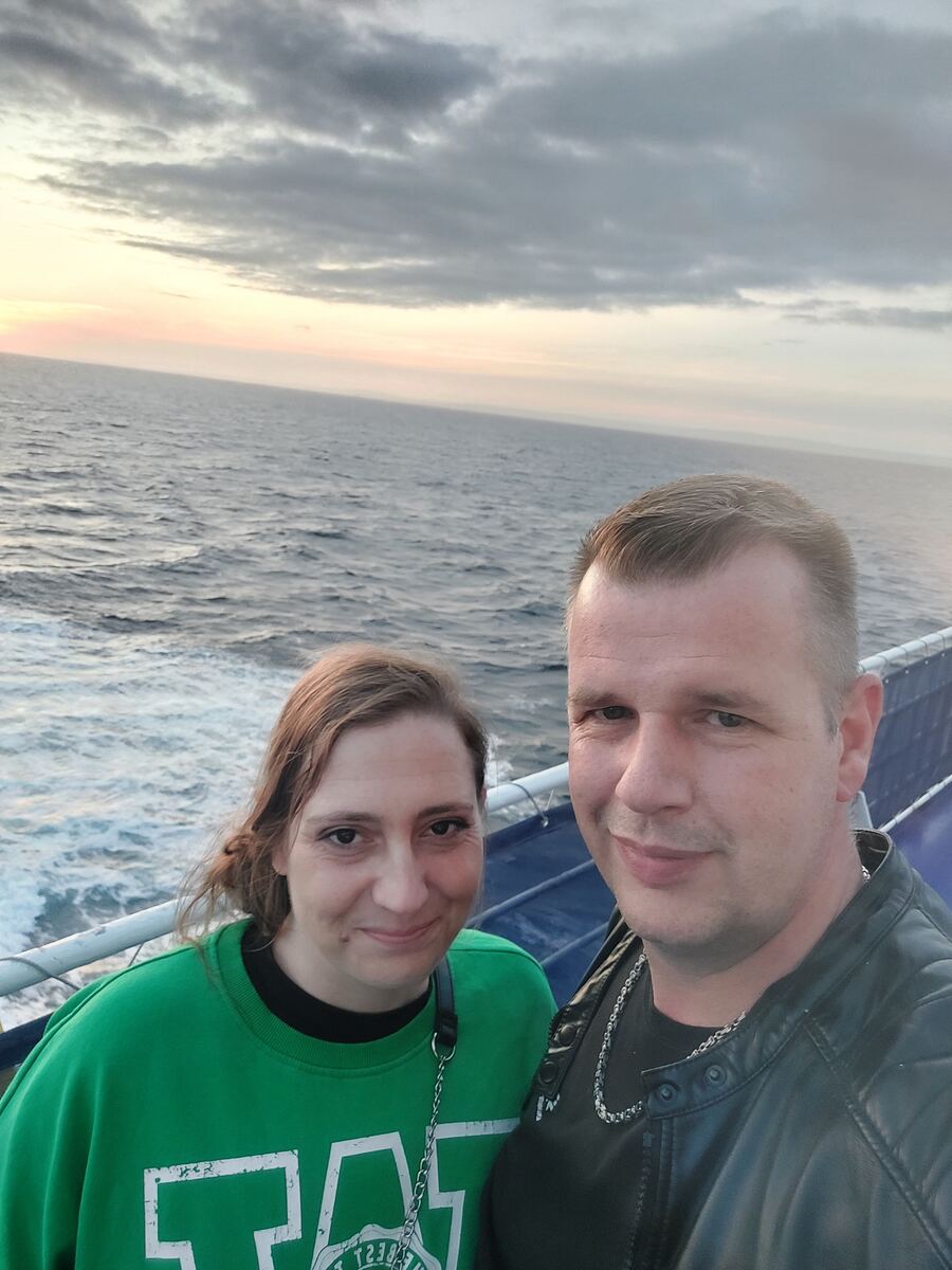 Een selfie van een stel op een boot of veerboot. Op de achtergrond is open zee te zien met lichte golven en de schuimende kielzog van het vaartuig. De lucht toont een zonsondergang met zachte oranje en roze tinten aan de horizon, overgaand in grijze wolken bovenin. De man draagt een donkere leren jas en de vrouw een groen sweatshirt. Ze staan bij een blauwe reling.