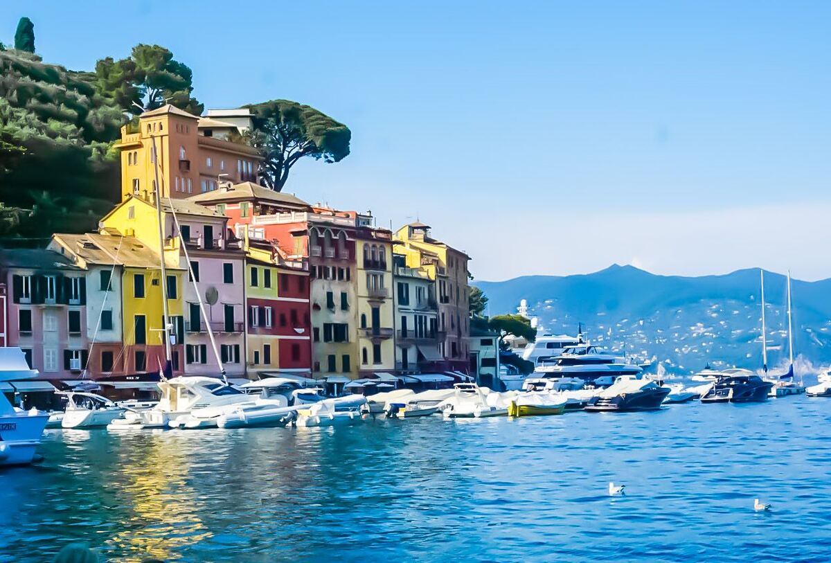 Een zonnige foto van de haven van Portofino met kleurrijke gebouwen langs de kustlijn en diverse boten in het water. Op de achtergrond zijn groene heuvels en bergen te zien onder een helderblauwe lucht.