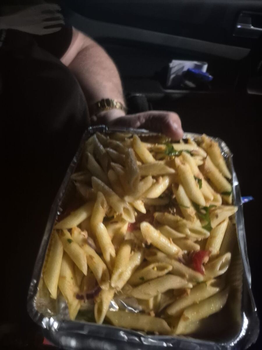 Een close-up van een aluminium afhaalbak met penne pasta, vermengd met stukjes tomaat, rode ui en groene kruiden. Een hand houdt de bak vast, en op de achtergrond is vaag het interieur van een auto te zien.