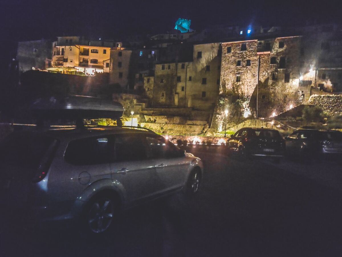 Een nachtfoto van een straat in Arcidosso, Italië, met een zilverkleurige Ford Focus stationwagen met dakkoffer op de voorgrond. Op de achtergrond zijn oude stenen gebouwen te zien die tegen een heuvel zijn gebouwd, verlicht door straatlantaarns. Bovenop de heuvel is een kasteel of fort te zien dat blauw is uitgelicht.