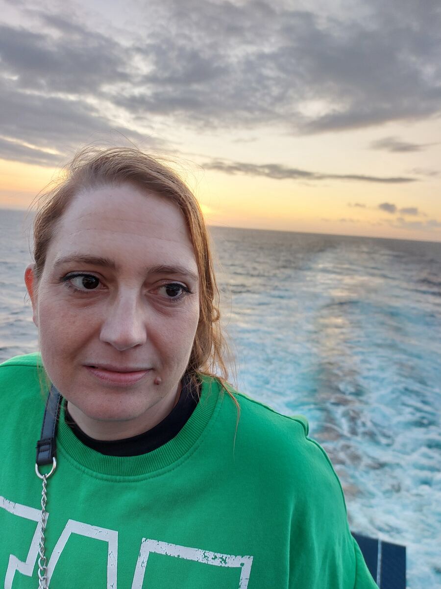 Een close-up van een vrouw in een groene trui, die naar rechtsboven kijkt. Achter haar is de open zee te zien met de golven die door een vaartuig worden gecreëerd. Aan de horizon is een zonsondergang zichtbaar met oranje en gele tinten, onder een deels bewolkte lucht.