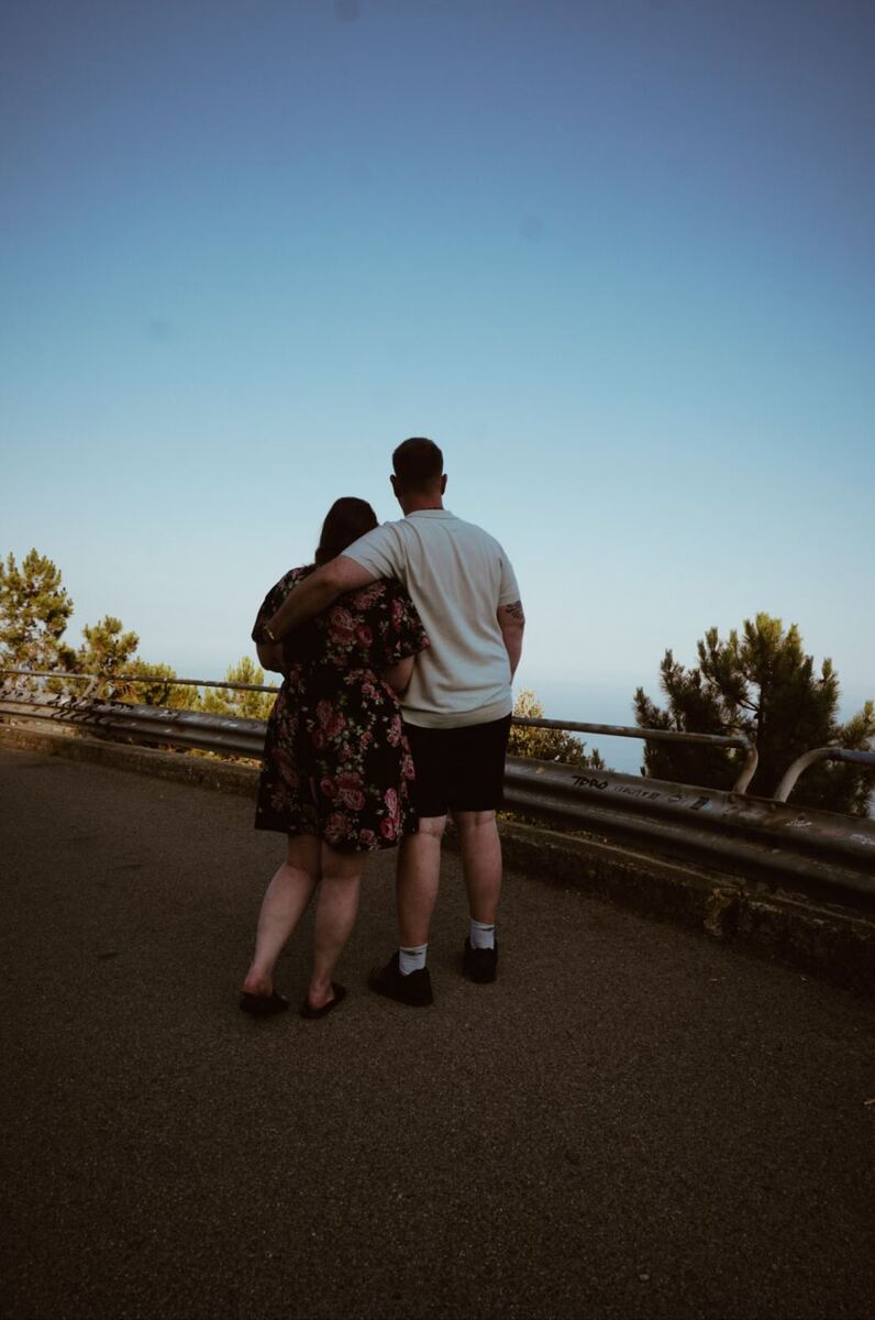 Een stel staat met de rug naar de camera, kijkend over een vangrail naar een landschap met bomen en de zee onder een heldere blauwe lucht. De vrouw draagt een donkere jurk met bloemenprint en de man een licht T-shirt met een donkere korte broek. Op de vangrail is graffiti zichtbaar.