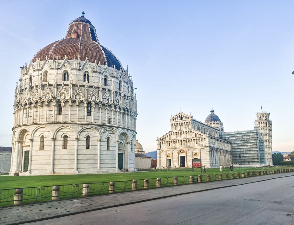 Een breed overzicht van de Piazza dei Miracoli in Pisa, Italië, met op de voorgrond een deel van een weg en grasveld. Links staat het ronde Baptisterium van Pisa met een koepeldak van rode dakpannen. Rechts daarvan bevindt zich de Kathedraal van Pisa, een groter wit marmeren gebouw met een donkere koepel. Achter de kathedraal is de Scheve Toren van Pisa zichtbaar, deels omgeven door steigers. De lucht is helderblauw.