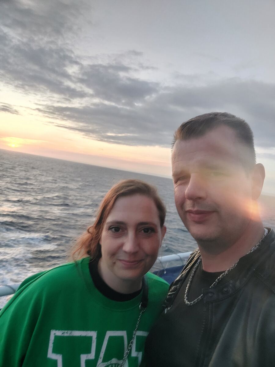 Een selfie van een man en een vrouw op een schip of veerboot, met de open zee en een zonsondergang op de achtergrond. De zon staat laag aan de horizon, wat zorgt voor warme oranje en roze tinten in de lucht, die overgaan in een bewolkte, grijze hemel. Het water is golvend en weerspiegelt het avondlicht. De man draagt een donker jack en een ketting, de vrouw een groen sweatshirt. Een deel van de reling van het schip is zichtbaar.