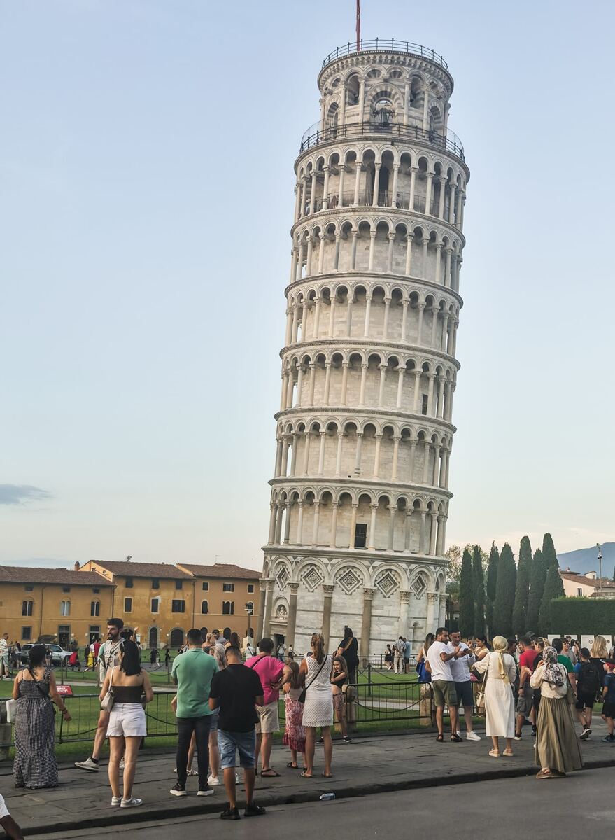 Een foto van de Scheve Toren van Pisa, een hoge, cilindervormige klokkentoren met meerdere galerijen van bogen en zuilen, die duidelijk overhelt. Rondom de toren is een openbaar plein met veel mensen, waaronder toeristen die foto's maken. Op de achtergrond zijn gebouwen met oranje daken en enkele hoge, slanke bomen zichtbaar. De lucht is lichtblauw en helder.