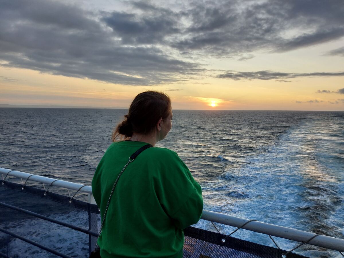Een persoon in een groene trui staat met de rug naar de camera gericht op een dek van een schip, kijkend naar de zonsondergang boven een kalme zee. De zon is laag aan de horizon en werpt een oranje gloed over het water en de lucht. Er zijn enkele wolken zichtbaar aan de hemel. Achter het schip is het schuimende kielzog te zien.
