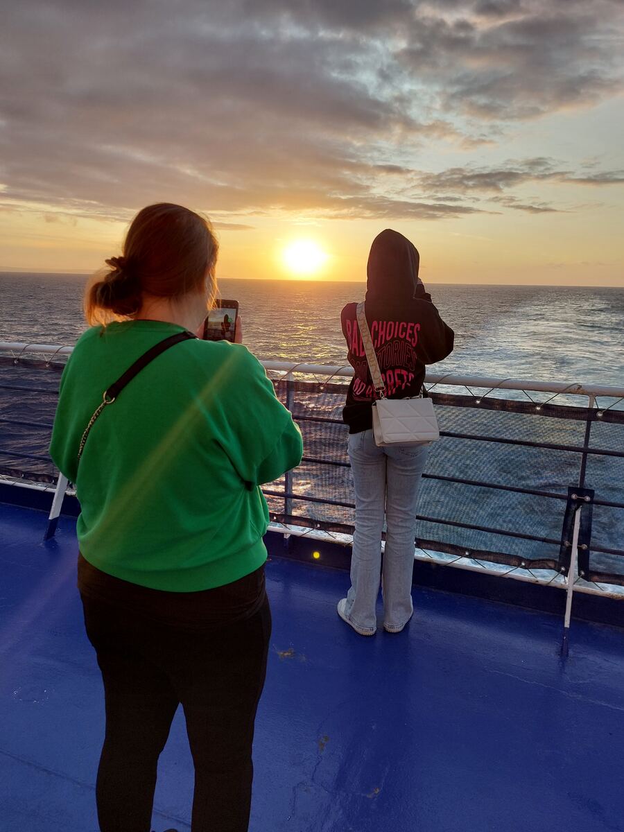 Twee personen staan op het dek van een veerboot en kijken naar de zonsondergang boven de zee. De zon is een heldere oranje bol aan de horizon, die reflecteert op het water. De lucht is deels bewolkt met oranje en grijze tinten. De persoon links, gekleed in een groen sweatshirt en zwarte broek, maakt een foto met een smartphone. De persoon rechts, gekleed in een donkere hoodie met roze tekst en een lichte spijkerbroek, kijkt ook naar de zonsondergang. Ze dragen een lichte schoudertas. Het dek van