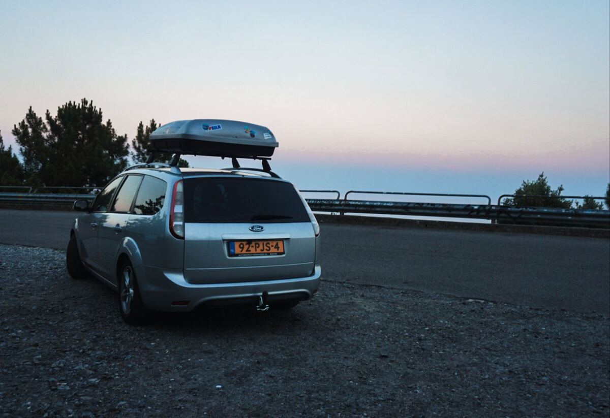 Een zilverkleurige Ford Focus stationwagen met een dakkoffer staat geparkeerd op een onverharde strook naast een geasfalteerde weg. Op de achtergrond is een hekwerk zichtbaar, met daarachter bomen en een wijds uitzicht over een landschap onder een avondlucht met zachte roze en blauwe tinten.