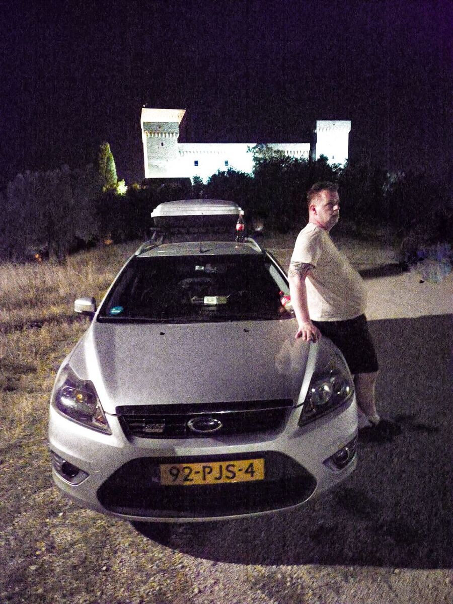 Een man leunt 's nachts tegen de motorkap van een zilverkleurige Ford Focus stationwagen met een dakkoffer. Op de achtergrond is een verlicht kasteel of fort te zien, omringd door donkere bomen en struiken. De lucht is donker en korrelig door de nachtopname.