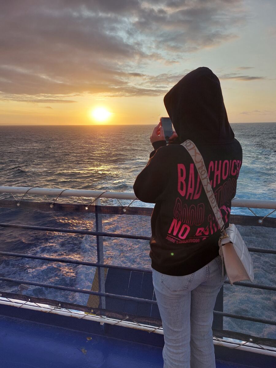 Een persoon in een zwarte hoodie met roze opdruk en een spijkerbroek staat met de rug naar de camera gericht op een dek van een schip. De persoon fotografeert met een smartphone de zonsondergang boven een kalme zee. De zon staat laag aan de horizon en werpt een gouden gloed over het water. Aan de horizon is een vage landmassa zichtbaar. De reling van het schip is op de voorgrond te zien.