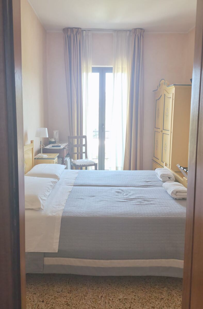 Een slaapkamer met twee naast elkaar geplaatste bedden, gedekt met een grijze sprei en witte lakens. Aan de rechterkant van het bed liggen twee opgerolde witte handdoeken. Links staat een nachtkastje met een lamp en een waterkoker. Een houten stoel met een gehaakt kleedje staat voor de ramen, die veel licht doorlaten en zijn voorzien van lichte gordijnen. Rechts in de kamer staat een lichtgekleurde kledingkast met decoratieve details. De muren zijn lichtroze en de vloer heeft een terrazzopatroon