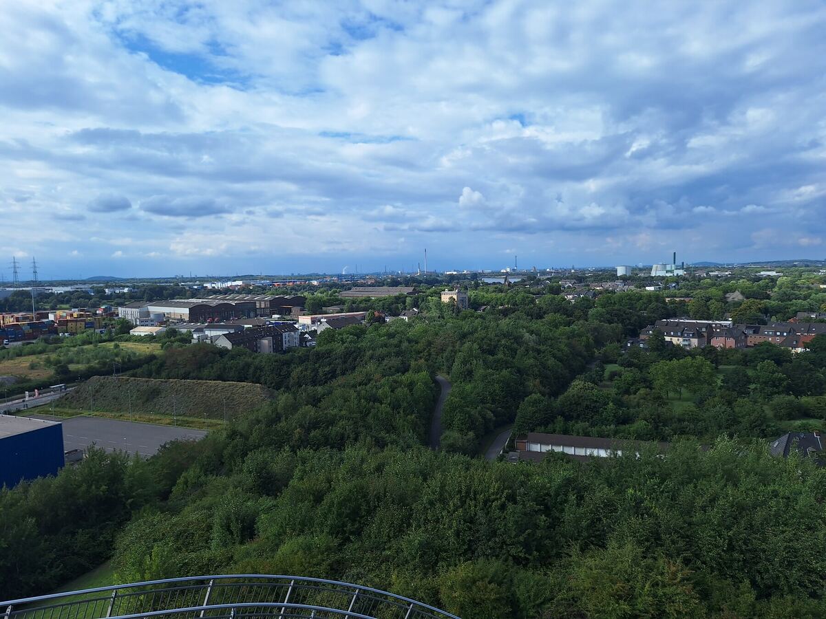 Een weids uitzicht over een stedelijk landschap met veel groen, industriële gebouwen en woonwijken onder een bewolkte lucht. Op de voorgrond is een deel van een metalen reling zichtbaar.
