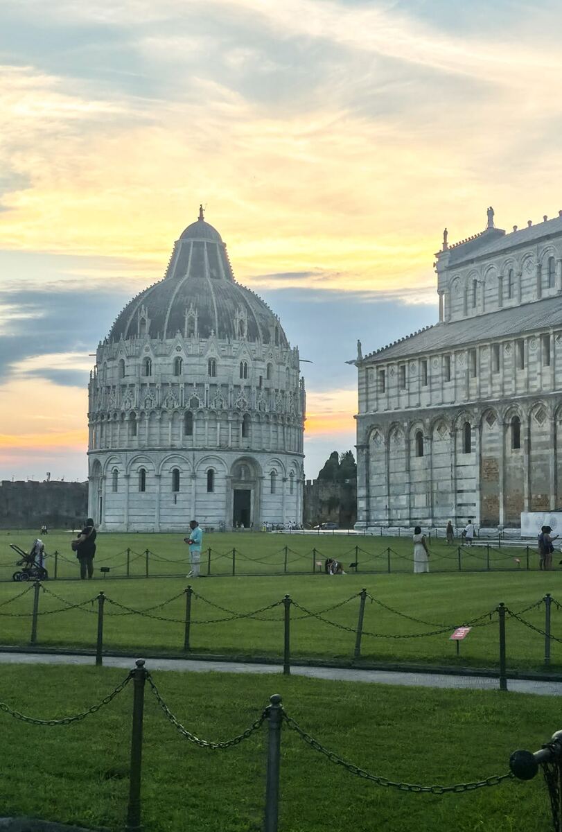 Een breed shot van de Piazza dei Miracoli in Pisa, Italië, met op de voorgrond een groen gazon afgebakend door een kettinghek. Links staat de ronde Battistero di San Giovanni met zijn kenmerkende koepel en gedetailleerde architectuur. Rechts is een deel van de Duomo di Pisa zichtbaar, eveneens in wit marmer met bogen en versieringen. Op het gazon staan enkele mensen verspreid. De lucht erboven toont een mix van oranje, gele en blauwe tinten, wat duidt op de late middag of vroege avond.