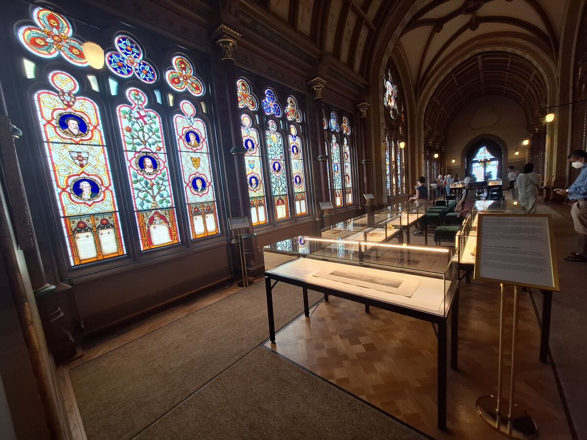 Een interieurfoto van een lange hal met aan de linkerkant een rij hoge, kleurrijke glas-in-loodramen. Voor de ramen staan vitrines met tentoongestelde voorwerpen. Aan de rechterkant van de hal is een gewelfd plafond zichtbaar en staan enkele bezoekers.