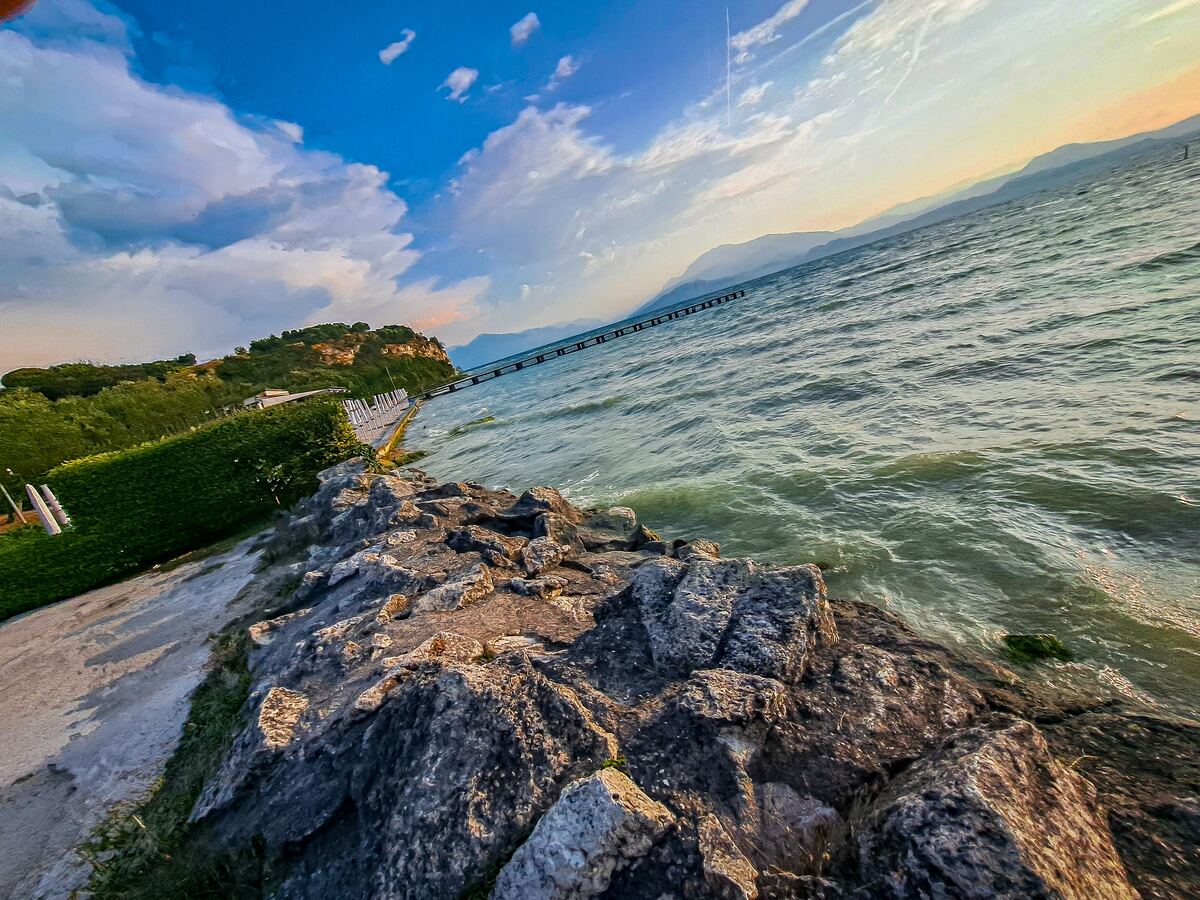 Een weids uitzicht over het Gardameer met rotsen op de voorgrond, een pier in de verte en bergen aan de horizon onder een deels bewolkte lucht.