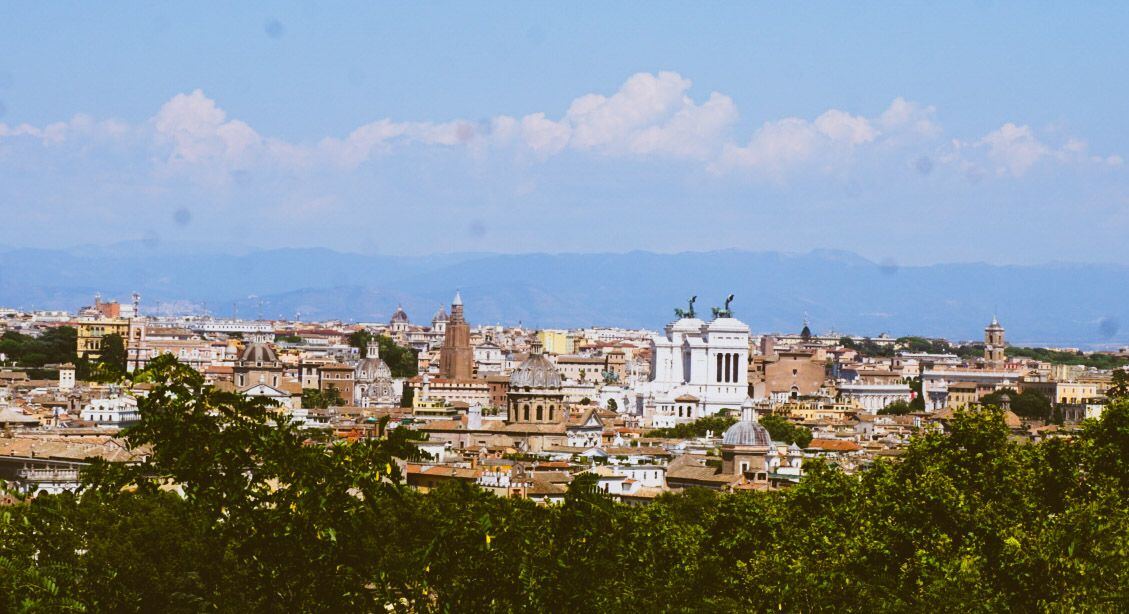 Een panoramisch uitzicht over de stad Rome met veel historische gebouwen, koepels en daken, gezien vanaf een hoger gelegen punt met bomen op de voorgrond. Op de achtergrond zijn bergen zichtbaar onder een lichtbewolkte blauwe lucht. Het Vittoriano is duidelijk herkenbaar in het midden van de foto.