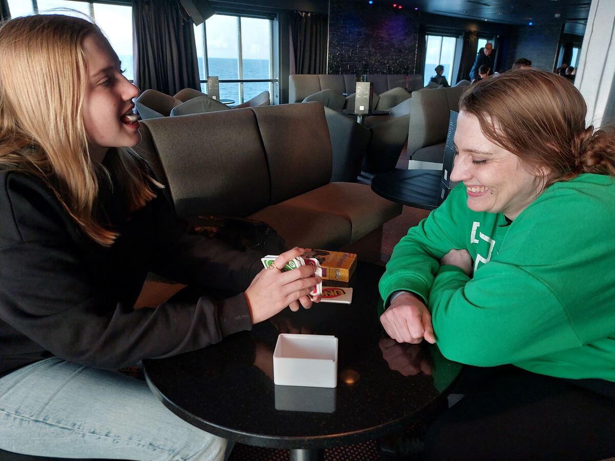 Twee vrouwen zitten aan een ronde tafel in een scheepsinterieur, met uitzicht op zee door grote ramen. De vrouw links, met lang blond haar en een zwarte trui, houdt speelkaarten vast en kijkt lachend naar de vrouw rechts. De vrouw rechts, met bruin haar in een knot en een groene trui, lacht ook en heeft haar armen over elkaar geslagen. Op tafel liggen een doosje Uno-kaarten en een wit bakje. Achter hen zijn comfortabele zitjes en tafels te zien, en in de verte andere passagiers.