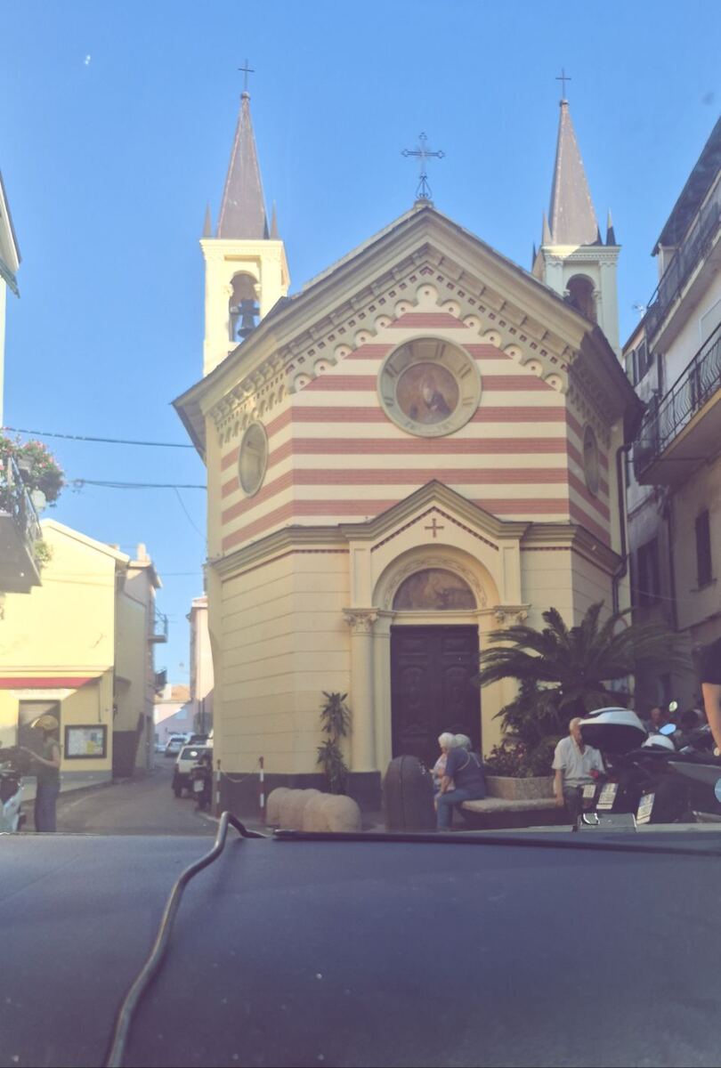 Een foto van een kerkgebouw met twee torens en een gestreepte gevel in rood en crème, gelegen aan een smalle straat. Voor de kerk zitten enkele mensen op een bankje en staan er scooters geparkeerd. De lucht is helderblauw.