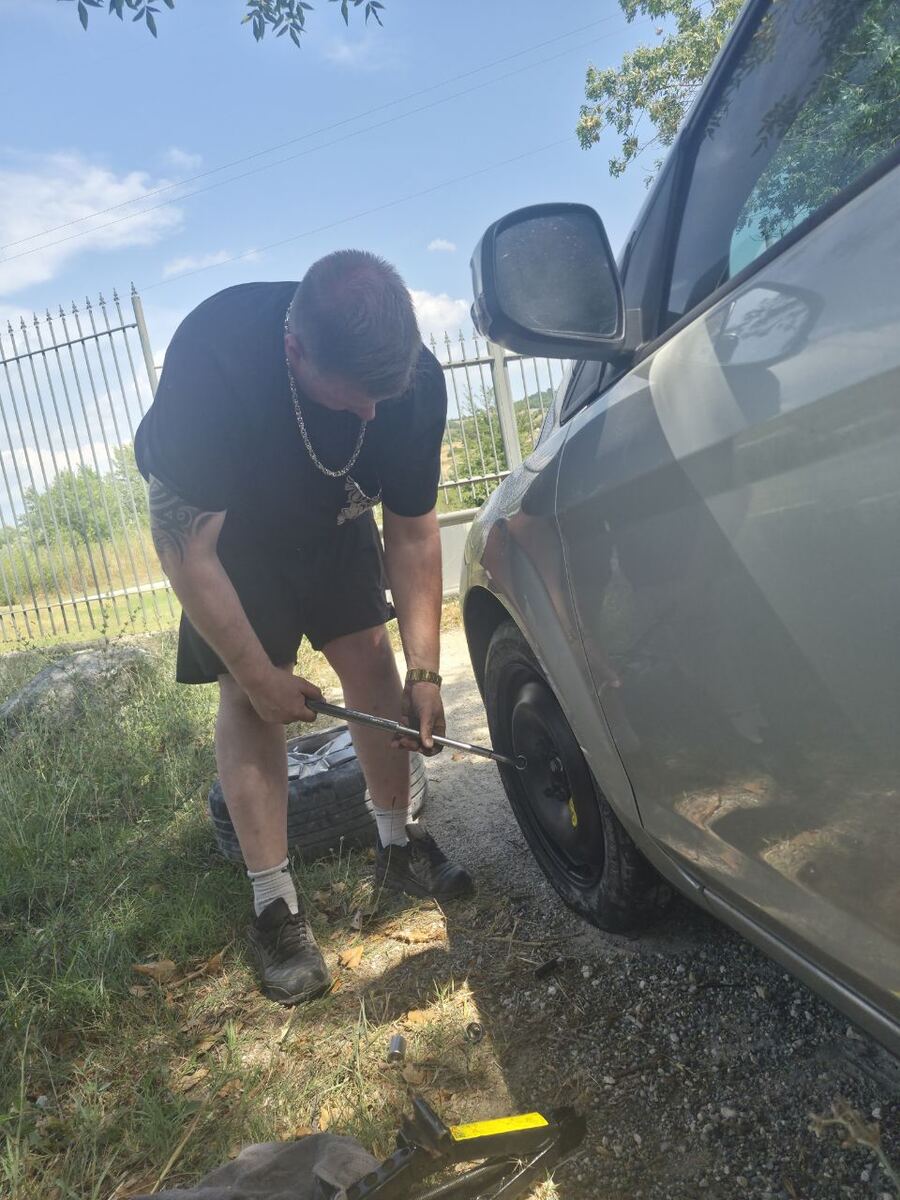 Een man in een zwart T-shirt en korte broek is bezig met het verwisselen van een autoband aan de zijkant van een zilverkleurige auto. Hij gebruikt een kruissleutel om de wielmoeren los te draaien. Op de grond liggen een krik en een reservewiel. De scène speelt zich af op een onverharde ondergrond met gras en grind, met op de achtergrond een metalen hek en groene begroeiing onder een helderblauwe lucht.