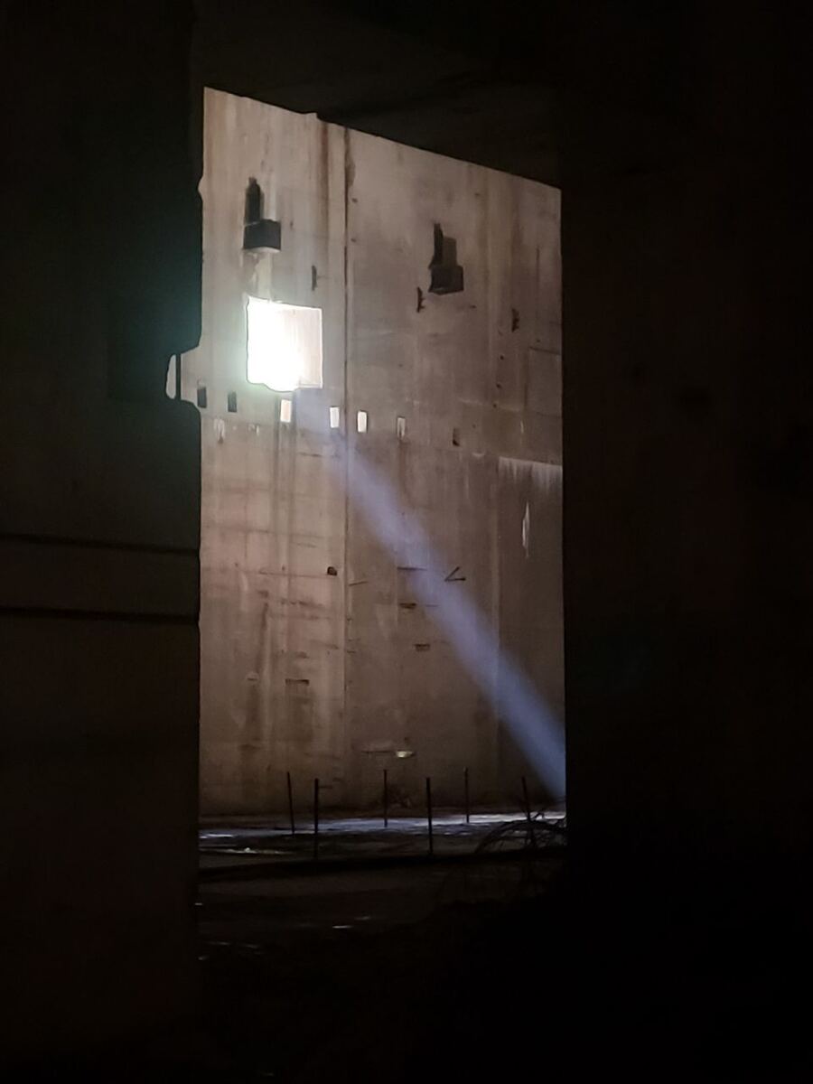 Een donkere, industri

le ruimte met een heldere lichtstraal die door een opening in de muur valt en de betonnen wanden verlicht. De muur heeft diverse kleine openingen en onregelmatigheden. De voorgrond is donker, waardoor de focus op de verlichte muur en lichtstraal ligt.