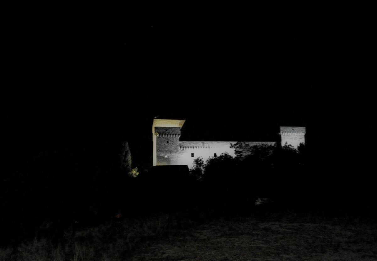 Een kasteel of fort is 's nachts verlicht, met een hoge toren aan de linkerkant en een lager gebouw ernaast. De rest van de omgeving is donker, met silhouetten van bomen en struiken op de voorgrond.