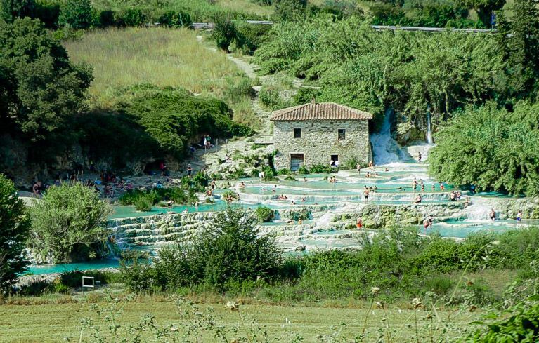 Een weids uitzicht op de Cascate del Mulino in Saturnia, Italië, met meerdere terrassen van turquoise water waarin mensen baden. Een stenen gebouw met een pannendak staat aan de rand van de baden, omringd door groene begroeiing en heuvels op de achtergrond. De zon schijnt helder op het landschap.