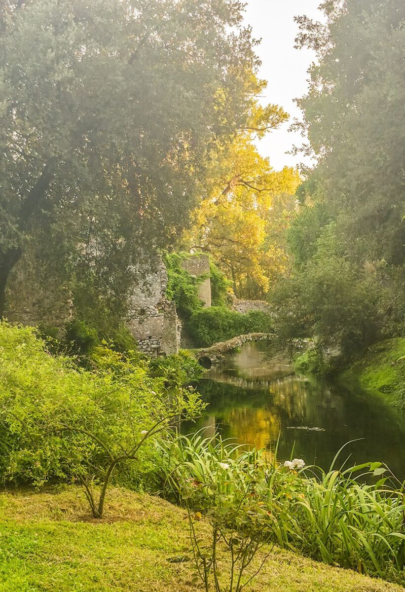 Een groene, bosrijke omgeving met een rustige waterloop in het midden. Aan de linkerkant staan de begroeide restanten van stenen muren en een ronde toren. Een kleine stenen brug, deels overwoekerd, verbindt de oevers over het water. De zon schijnt door de bladeren van de bomen en creëert een lichte gloed op het wateroppervlak.