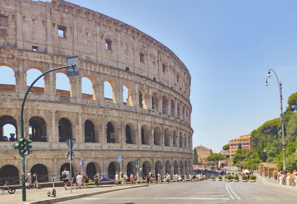Een zonnige foto van het Colosseum in Rome, gezien vanaf de straat. De imposante structuur van het amfitheater domineert de linkerkant van het beeld, met zijn karakteristieke bogen en verdiepingen. Op de voorgrond is een brede weg te zien met verkeerslichten en een aantal voetgangers op de stoep. Aan de rechterkant van de weg zijn bomen en gebouwen zichtbaar onder een helderblauwe lucht.