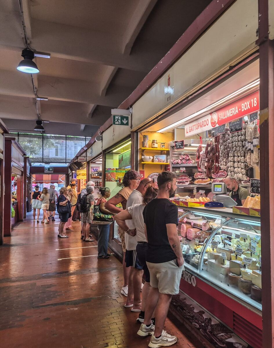 Een binnenmarkt met een lange rij mensen die voor een salumeria staan. De kraam toont een vitrine vol kazen en diverse vleeswaren, met daarboven hangende worsten en hammen. Achter de toonbank staat een verkoper. De vloer is van rode tegels en het plafond heeft industriële lampen en betonnen balken. Er is een groene nooduitgangaanduiding zichtbaar.