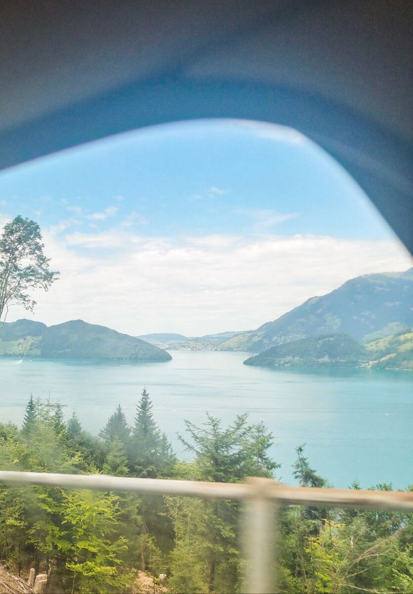 Een foto genomen vanuit een voertuig, met een deel van het dak en de raamstijl zichtbaar. We zien een breed uitzicht over een groot meer met aan weerszijden groene, beboste bergen. Aan de overkant van het meer is een kleine nederzetting te zien. Op het water vaart een boot en op de voorgrond zijn naaldbomen en een metalen reling zichtbaar.