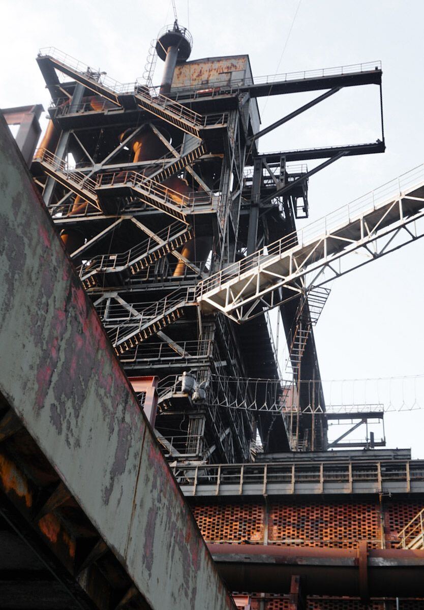 Een close-up van een groot industrieel complex met veel stalen constructies, trappen en bruggen, deels roestig en verweerd. De lucht is licht en helder, wat de details van de constructie goed laat uitkomen.
