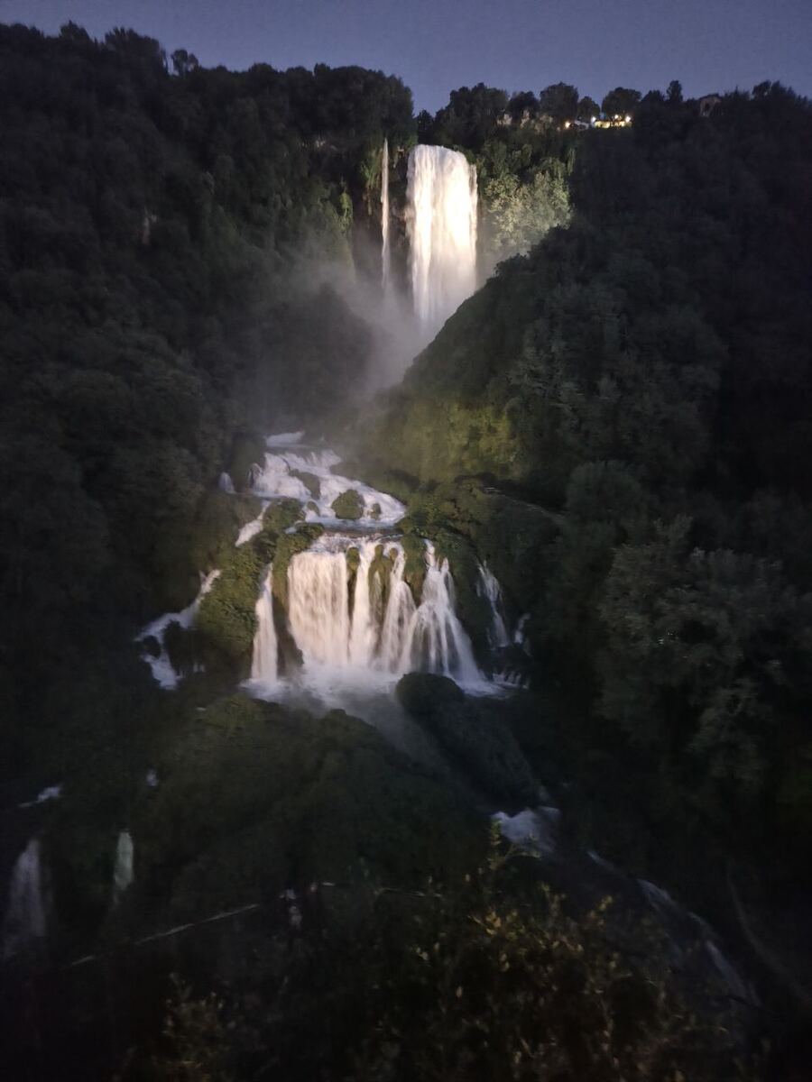 Een verticale foto van een verlichte waterval in de avond, omringd door donkere, dichtbegroeide heuvels. Het water valt in meerdere etappes naar beneden, waarbij de bovenste val het meest prominent is en fel wordt verlicht. De lucht boven de heuvels is donkerblauw, wat duidt op schemering of nacht. Er zijn enkele lichtjes zichtbaar op de top van de heuvels.