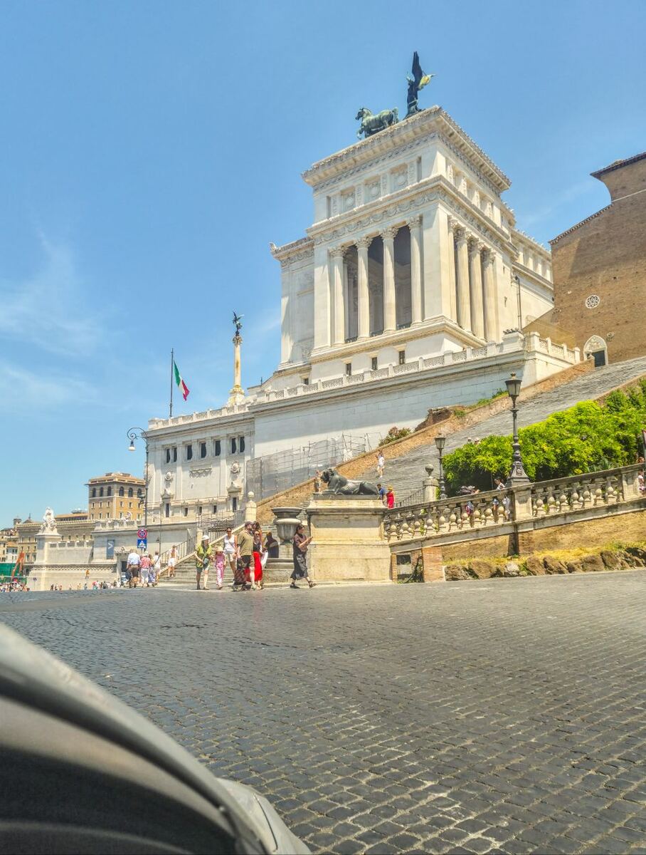 Een foto genomen vanaf straatniveau met een deel van een auto op de voorgrond, met op de achtergrond het grote witte Monument van Victor Emanuel II in Rome onder een helderblauwe lucht. Mensen lopen op de geplaveide straat en de trappen van het monument. De Italiaanse vlag wappert bij het gebouw.