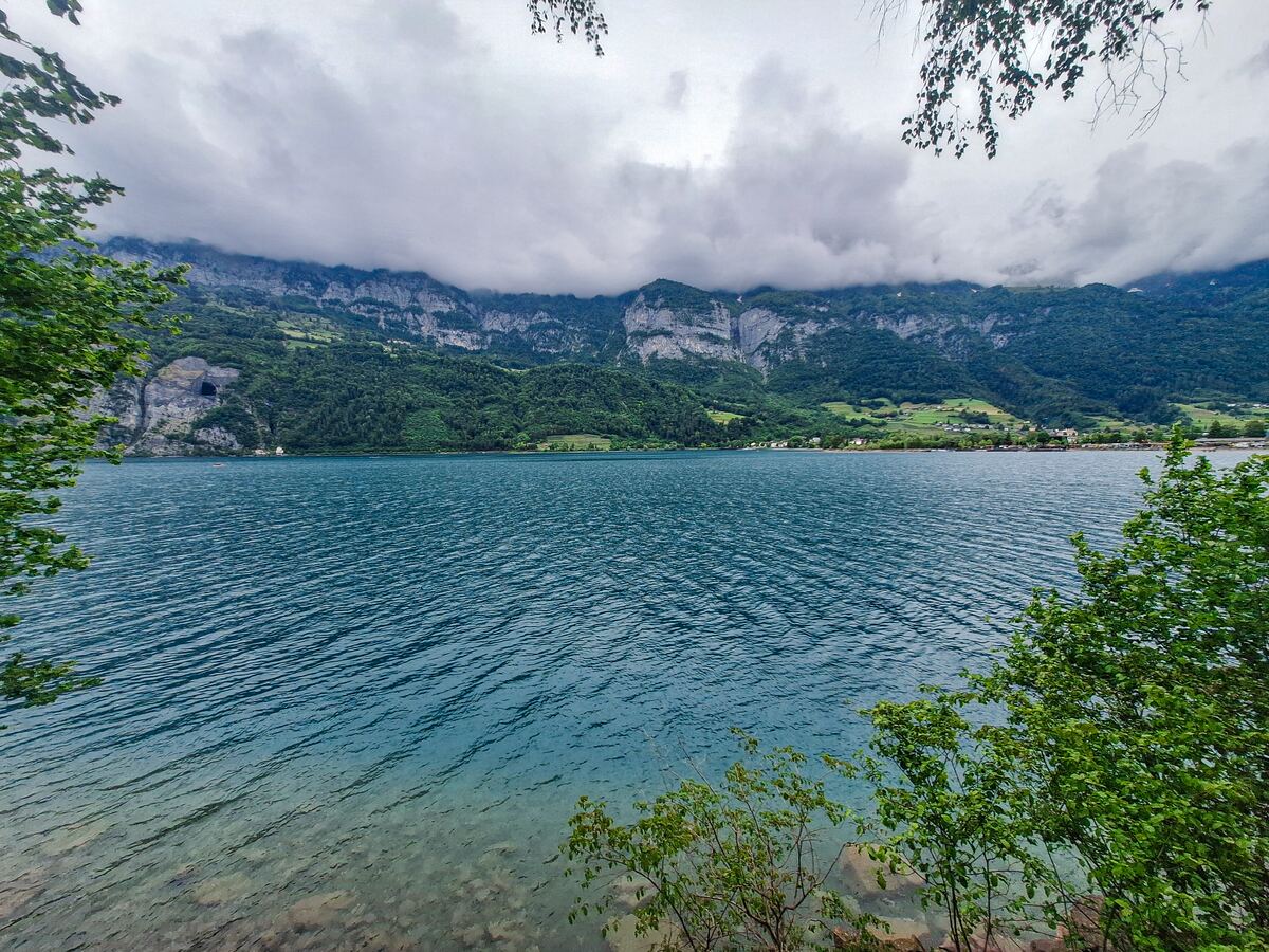 Een breedbeeldopname van een groot meer met turquoise water, omringd door groene bergen. De lucht is bewolkt en de bergen zijn deels bedekt met wolken. Op de voorgrond zijn groene bladeren van bomen zichtbaar die het beeld aan de boven- en onderkant omlijsten. Aan de overkant van het meer is een kleine nederzetting te zien aan de voet van de bergen.