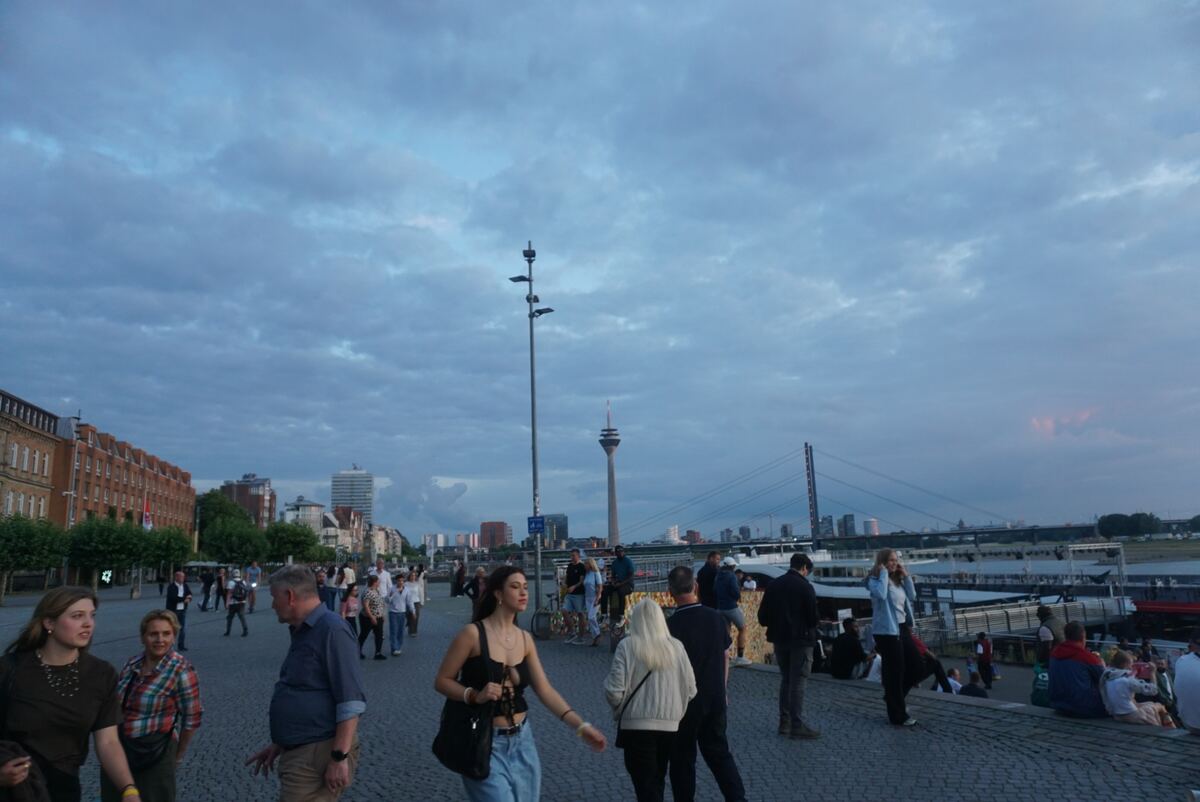 Een avondbeeld van een drukke promenade langs een rivier, met mensen die wandelen en zitten. Op de achtergrond zijn gebouwen, een brug en een televisietoren zichtbaar onder een bewolkte lucht.