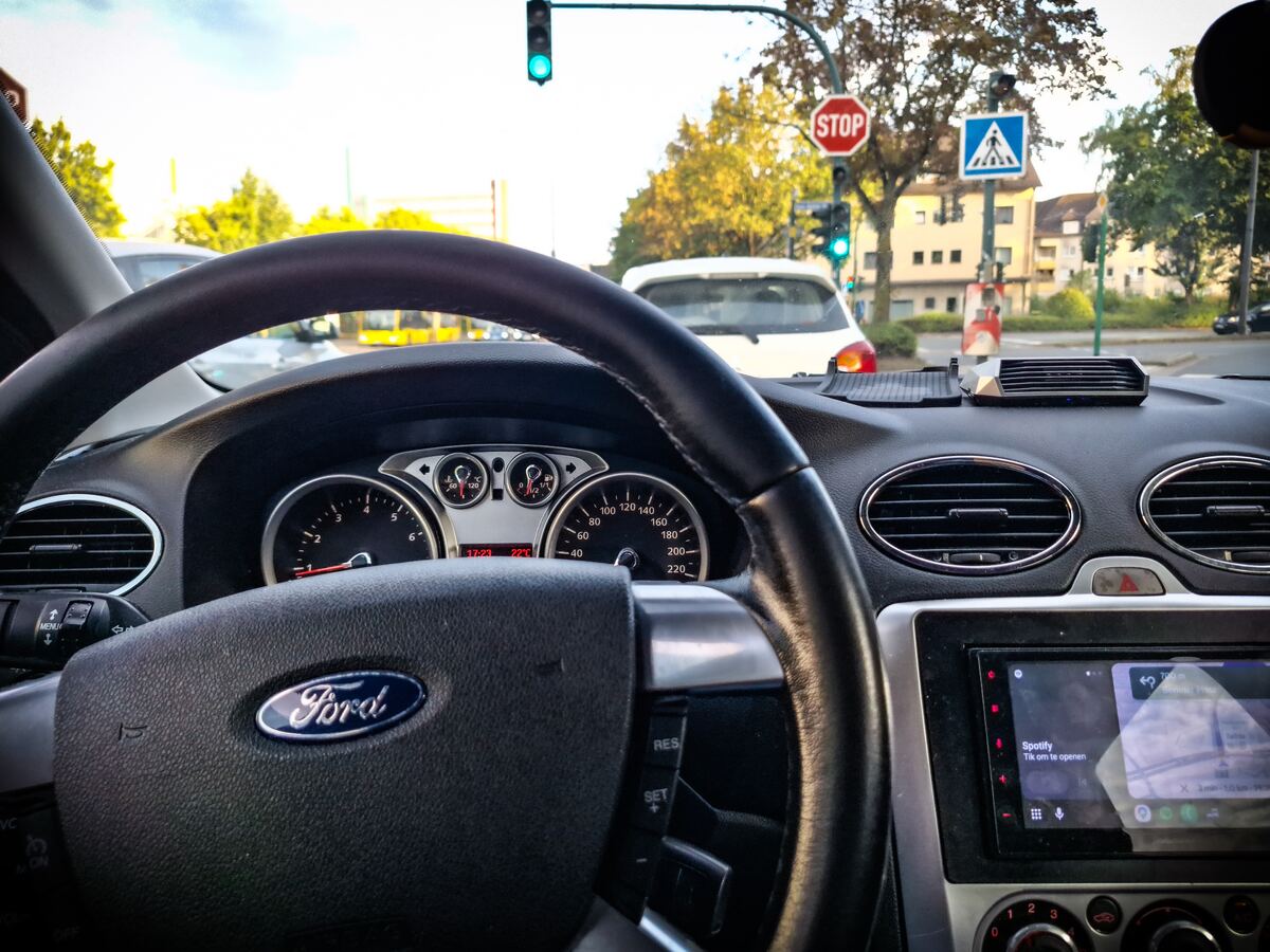 Een interieurfoto vanuit een auto, met het stuur en het dashboard op de voorgrond. Door de voorruit is een kruispunt te zien met een groen verkeerslicht, een stopbord en een zebrapadbord. De zon staat laag en schijnt fel.