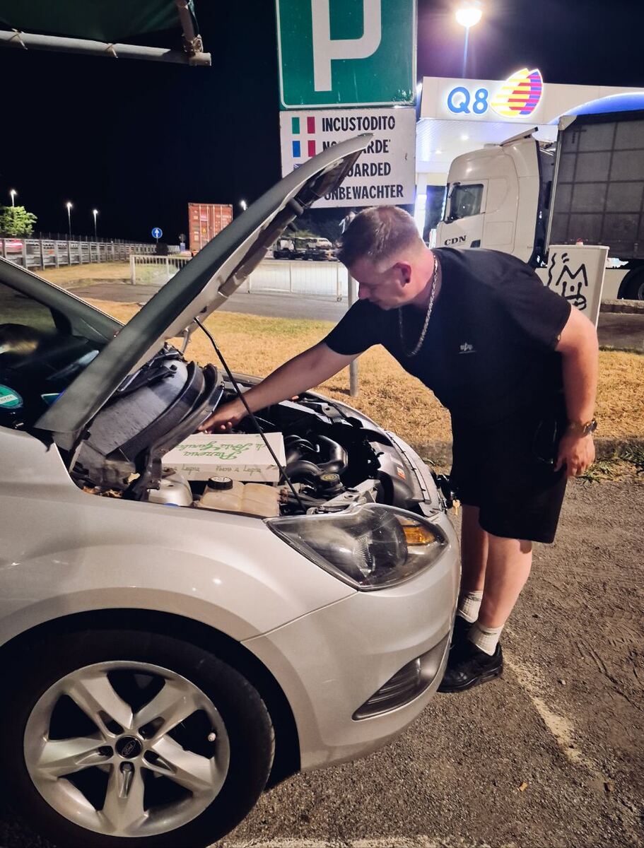 Een man staat 's nachts bij de open motorkap van een zilverkleurige Ford Focus stationwagon op een parkeerplaats. Op de achtergrond is een groen parkeerbord te zien met de tekst 'INCUSTODITO' en vlaggen van Italië en Frankrijk, en een Q8 tankstationlogo.
