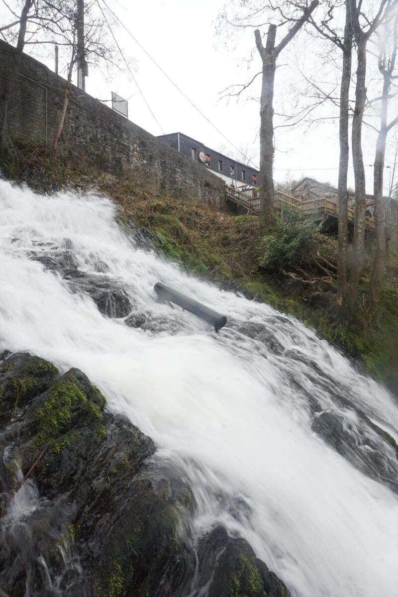 Een waterval stroomt over rotsen met mos, met op de achtergrond een stenen muur, kale bomen en een gebouw met een houten trap.