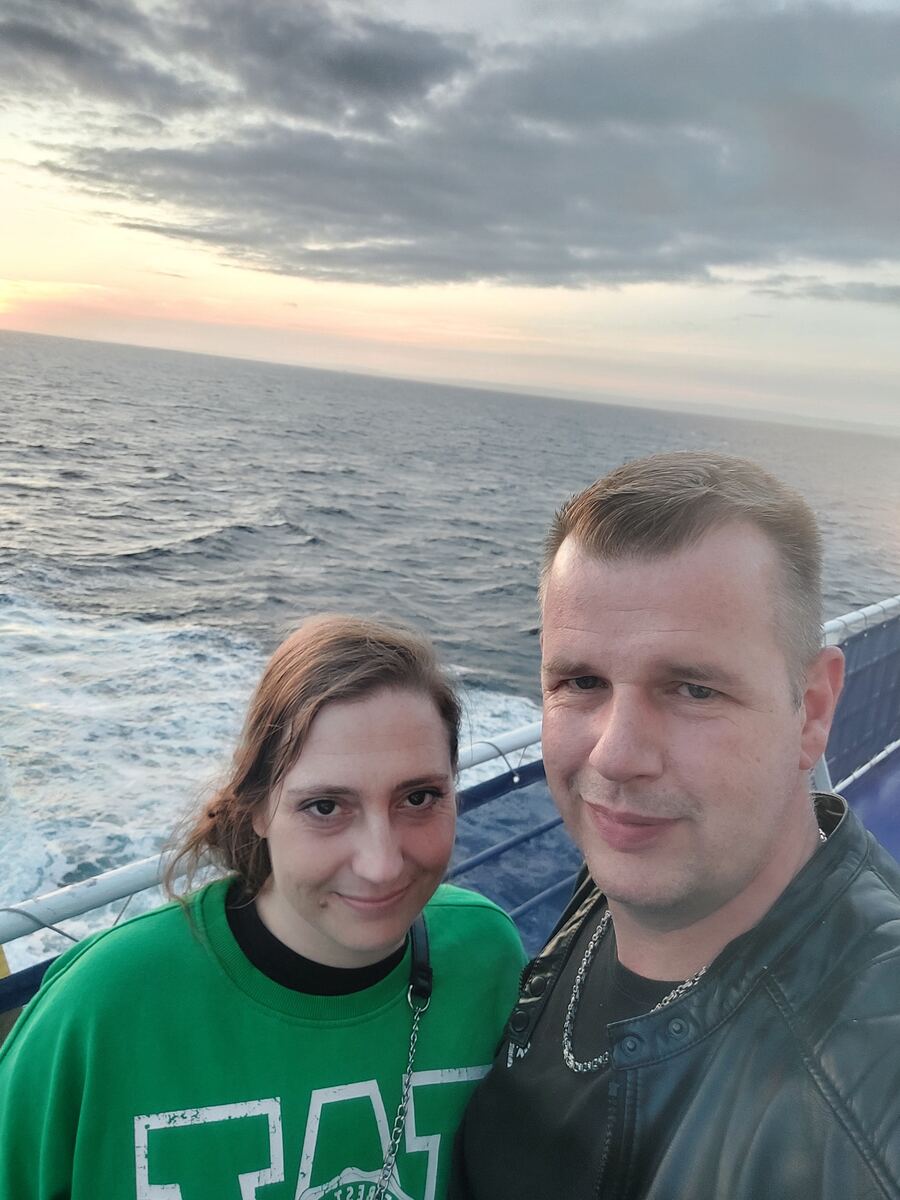 Een selfie van een stel op het dek van een veerboot, met de zee en een zonsondergang op de achtergrond. De vrouw draagt een groene trui en de man een zwart T-shirt met een leren jasje.