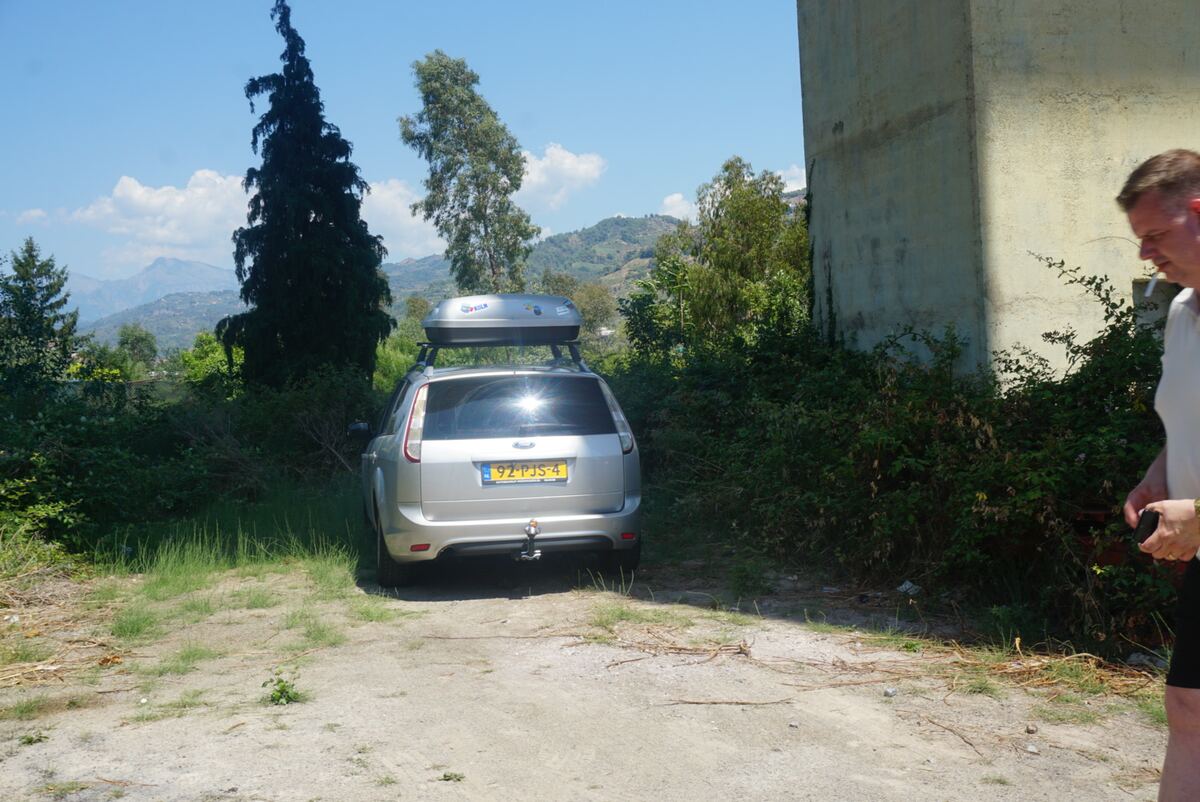 Een zilverkleurige Ford Focus stationwagen met een dakkoffer staat geparkeerd op een onverharde ondergrond naast een betonnen muur en begroeiing. Op de achtergrond zijn bergen en bomen te zien onder een blauwe lucht met enkele wolken. Rechts in beeld is een deel van een persoon zichtbaar.