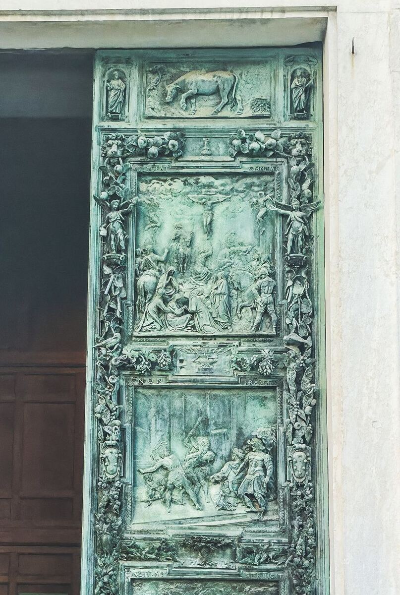 Een close-up van een grote, groen uitgeslagen bronzen deur met gedetailleerde reliëfs. De deur toont verschillende scènes, waaronder een kruisiging en andere figuren, omringd door decoratieve elementen zoals bladeren en kleine sculpturen. Rechts van de deur is een lichte stenen muur zichtbaar.