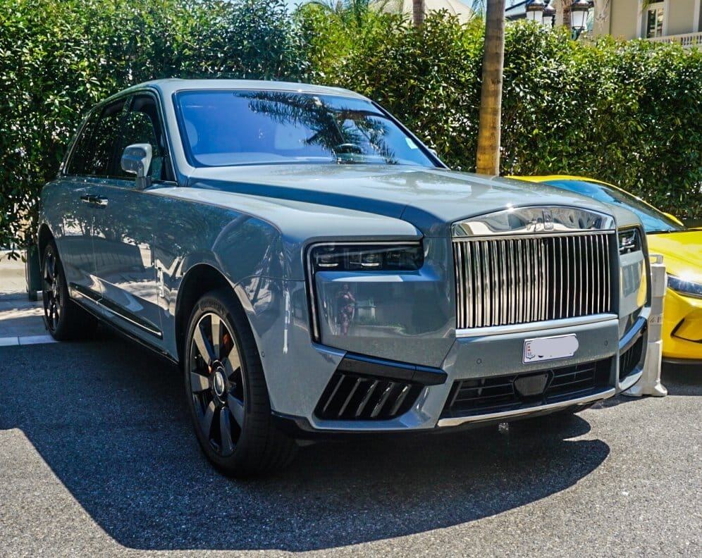 Een lichtgrijze Rolls-Royce Cullinan staat geparkeerd op een asfaltweg onder fel zonlicht. Rechtsachter is een deel van een gele sportwagen zichtbaar. De achtergrond bestaat uit groene heggen en de stam van een palmboom, met een gebouw in de verte.