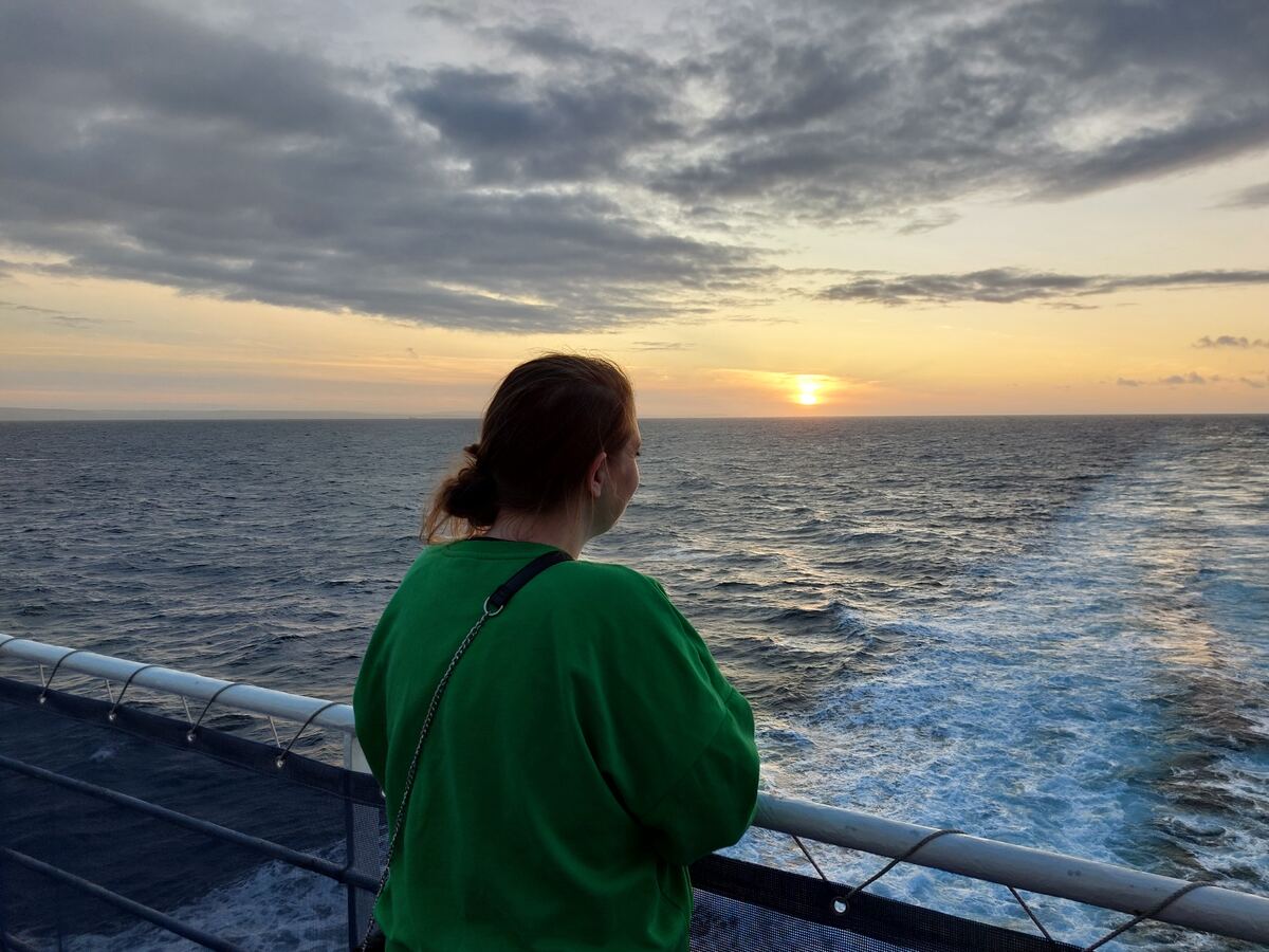 Een persoon in een groen shirt kijkt uit over de zee waar de zon ondergaat. De lucht is deels bewolkt met oranje en grijze tinten. De persoon staat bij een reling van wat een schip lijkt te zijn, met schuimend water achter het schip.