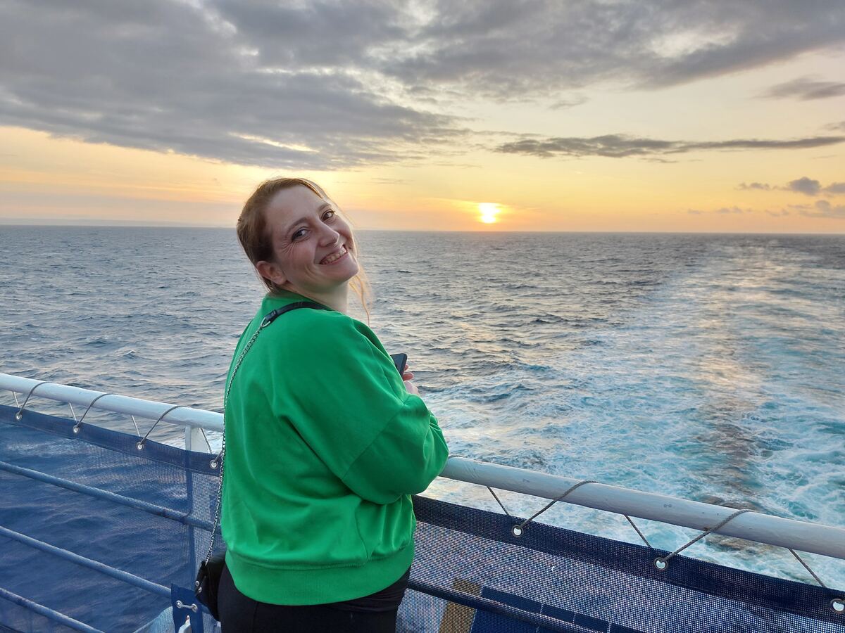 Een vrouw in een groene trui staat op het dek van een schip en kijkt lachend over haar schouder naar de camera. Achter haar is de open zee te zien met een zonsondergang aan de horizon. De zon is een heldere stip aan de hemel, die de wolken en het water in warme tinten kleurt. Het schip laat een schuimend kielzog achter in het water.