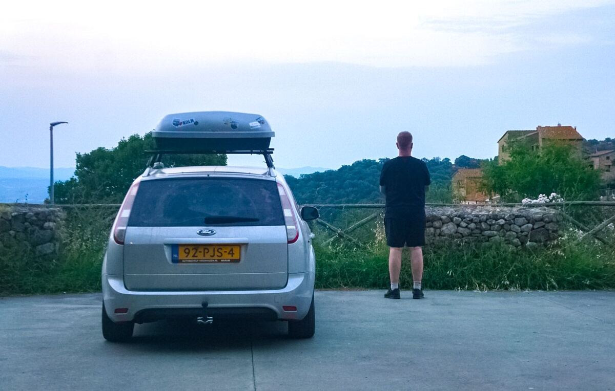 Een man staat met zijn rug naar de camera en kijkt uit over een heuvelachtig landschap met bebouwing in de verte. Links staat een zilverkleurige Ford Focus stationwagen met een dakkoffer. De lucht is lichtblauw met wat wolken. Op de achtergrond zijn groene bomen en struiken te zien, en enkele huizen met oranje daken.