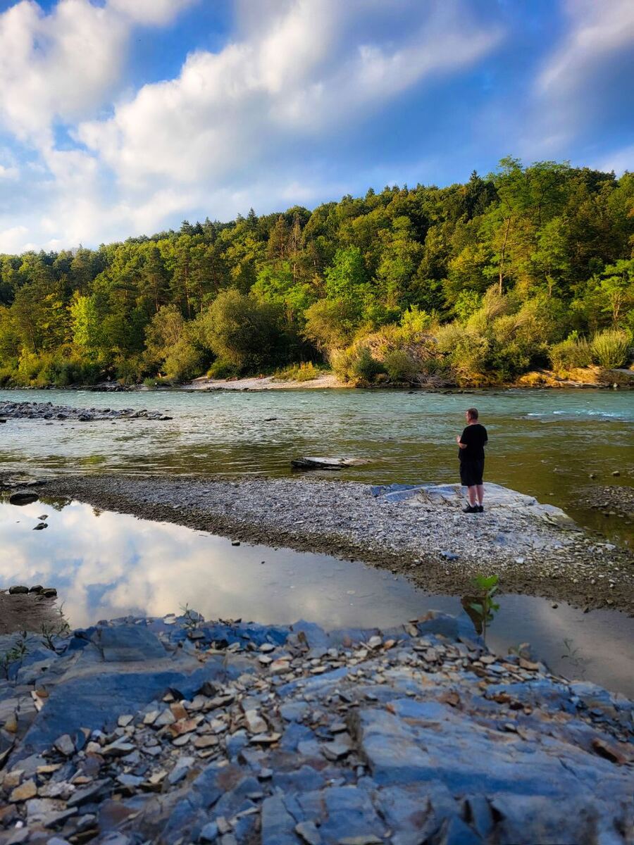 Een persoon staat op een smalle strook land in een rivier, omringd door water. Op de achtergrond is een dichtbegroeide groene heuvel te zien onder een blauwe lucht met witte wolken. De voorgrond bestaat uit rotsen en kiezels.