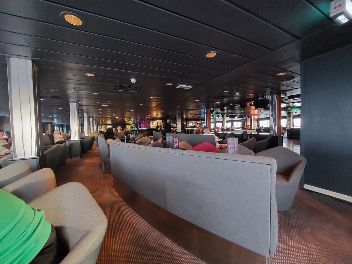 Een ruime lounge met een donker plafond en rijen grijze zitplaatsen. Mensen zitten verspreid in de ruimte, sommigen bij een bar achterin. Aan de rechterkant zijn ramen zichtbaar die daglicht doorlaten.