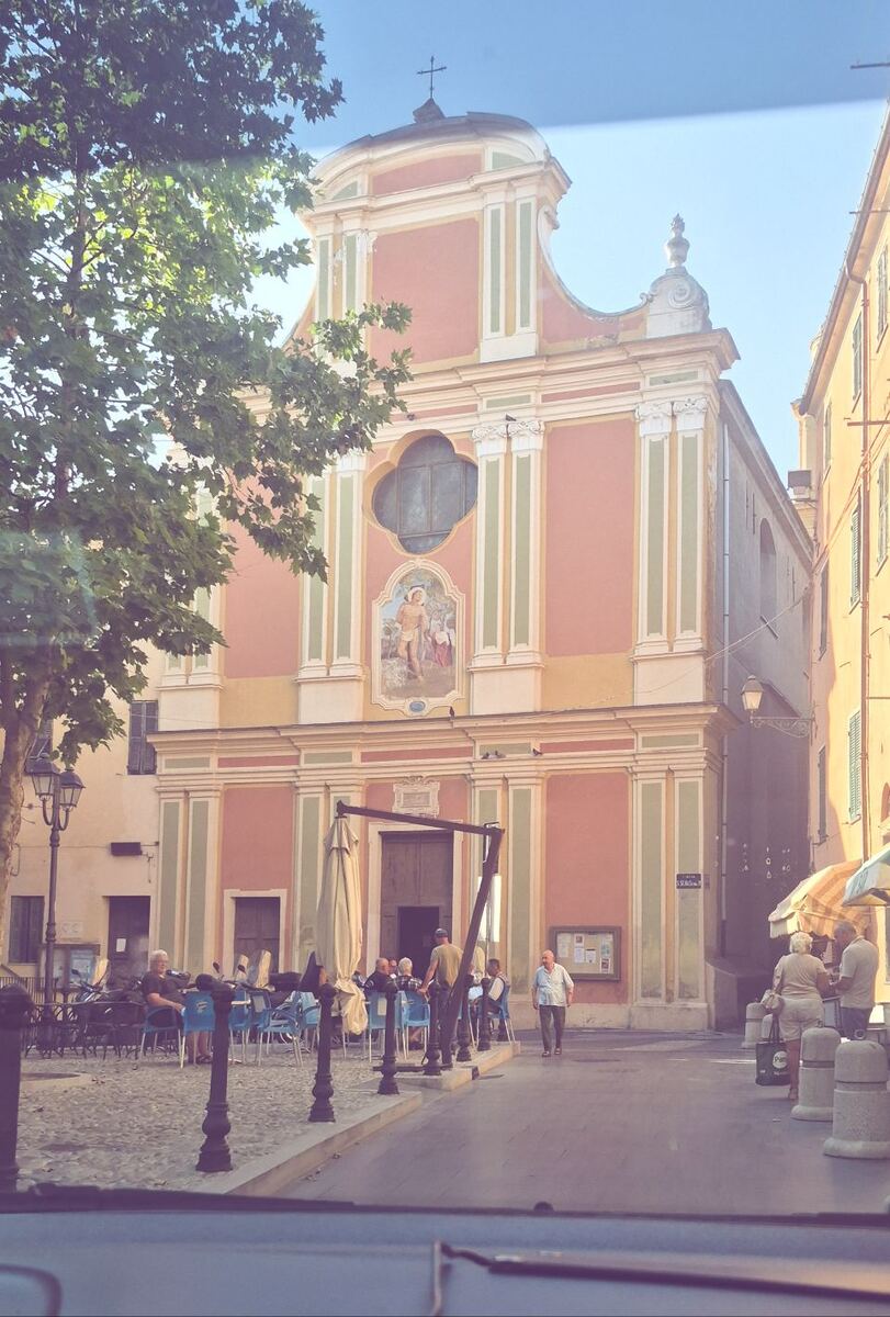 Een foto van een roze-gele kerk met een fresco boven de ingang, gelegen aan een plein met terrassen en mensen. De foto is genomen vanuit een auto, met een deel van de voorruit zichtbaar aan de onderkant.