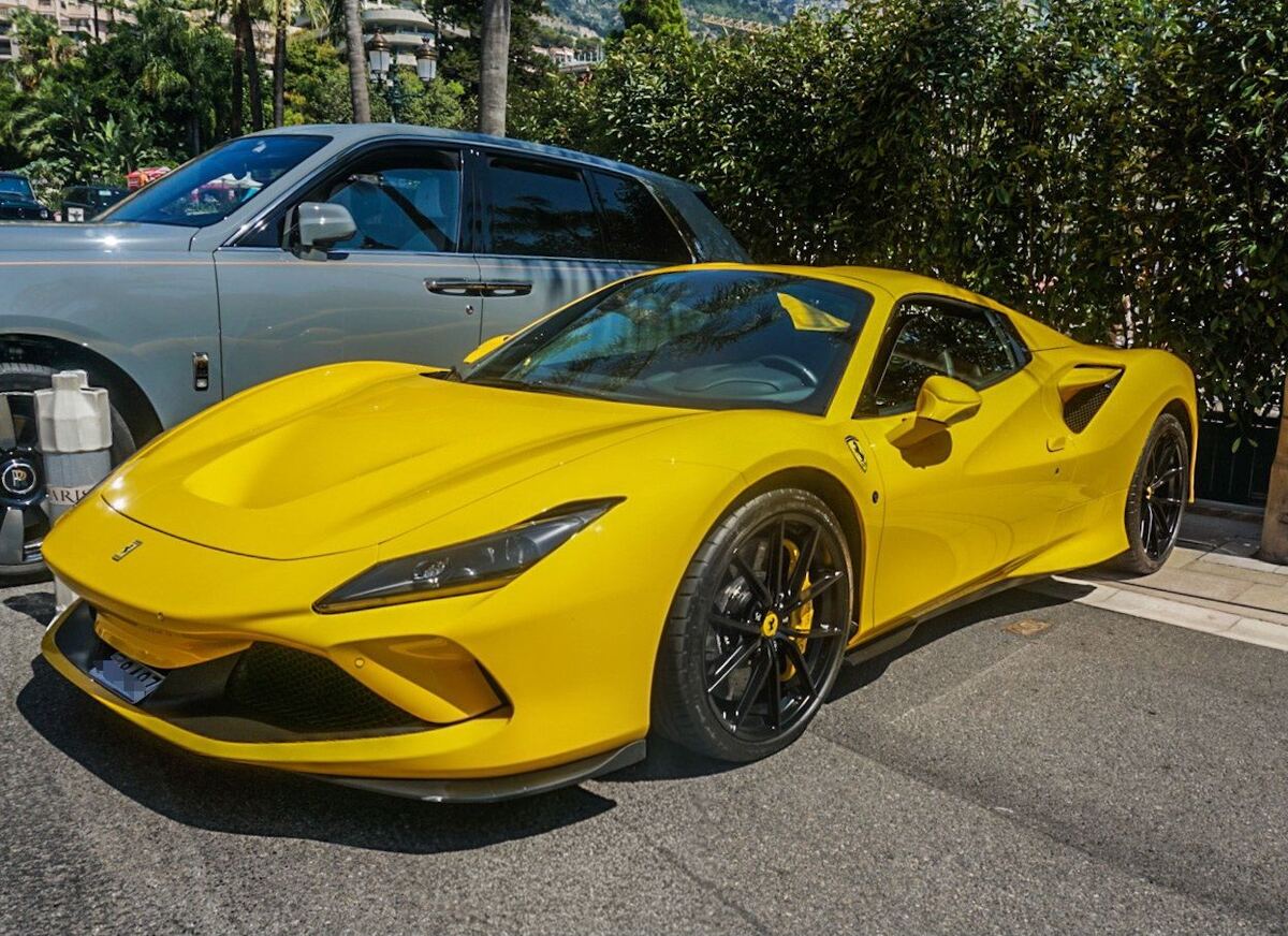 Een felgele Ferrari F8 Tributo staat geparkeerd op een zonnige straat. Achter de Ferrari is een grijze Rolls-Royce Cullinan deels zichtbaar. Op de achtergrond zijn groene struiken, palmbomen en gebouwen op een heuvel te zien.