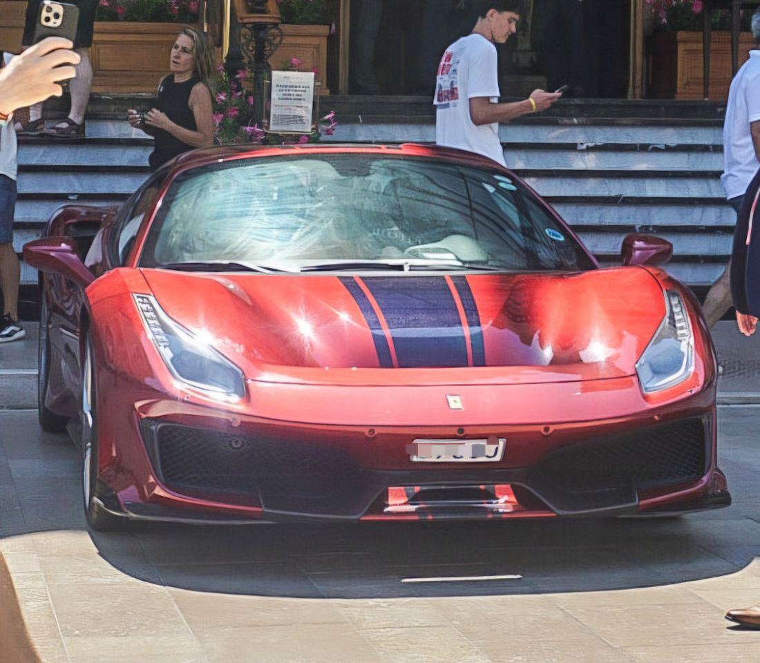 Een rode sportauto met donkere strepen over de motorkap staat geparkeerd voor een gebouw met een trap. Op de achtergrond zijn mensen te zien, waarvan één een foto maakt met een smartphone.